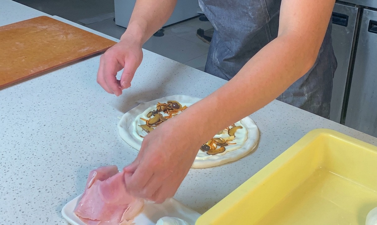 Mani di uno chef che sfilacciano fette di prosciutto cotto sopra una pizza condita con funghi e salsa bianca.
