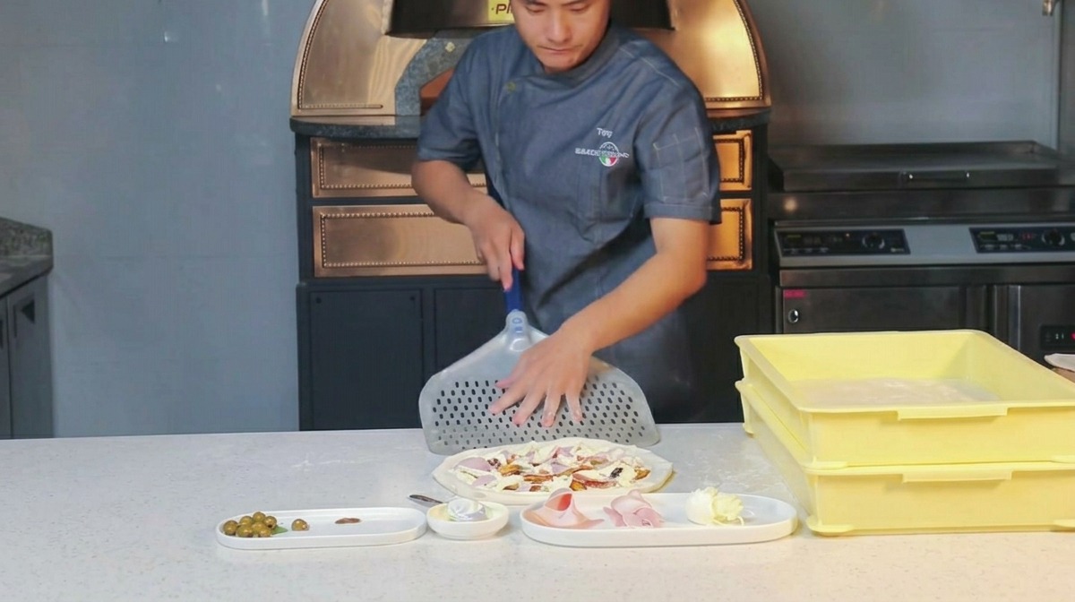 Uno chef che sfrega della farina su una pala per pizza in metallo forata.