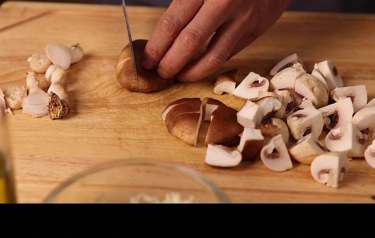 Mani che affettano funghi shiitake e champignon freschi su un tagliere di legno.