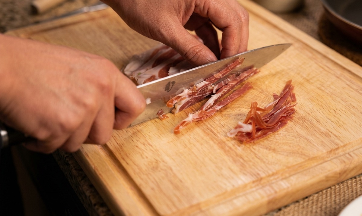 Mani che tagliano a strisce sottili del prosciutto crudo con un coltello da chef.