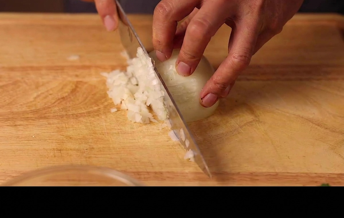 Mani che tritano finemente mezza cipolla bianca su un tagliere di legno.