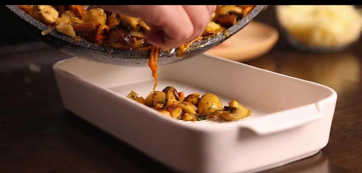 Padella che versa il mix di funghi cotti in una teglia da forno in ceramica bianca.