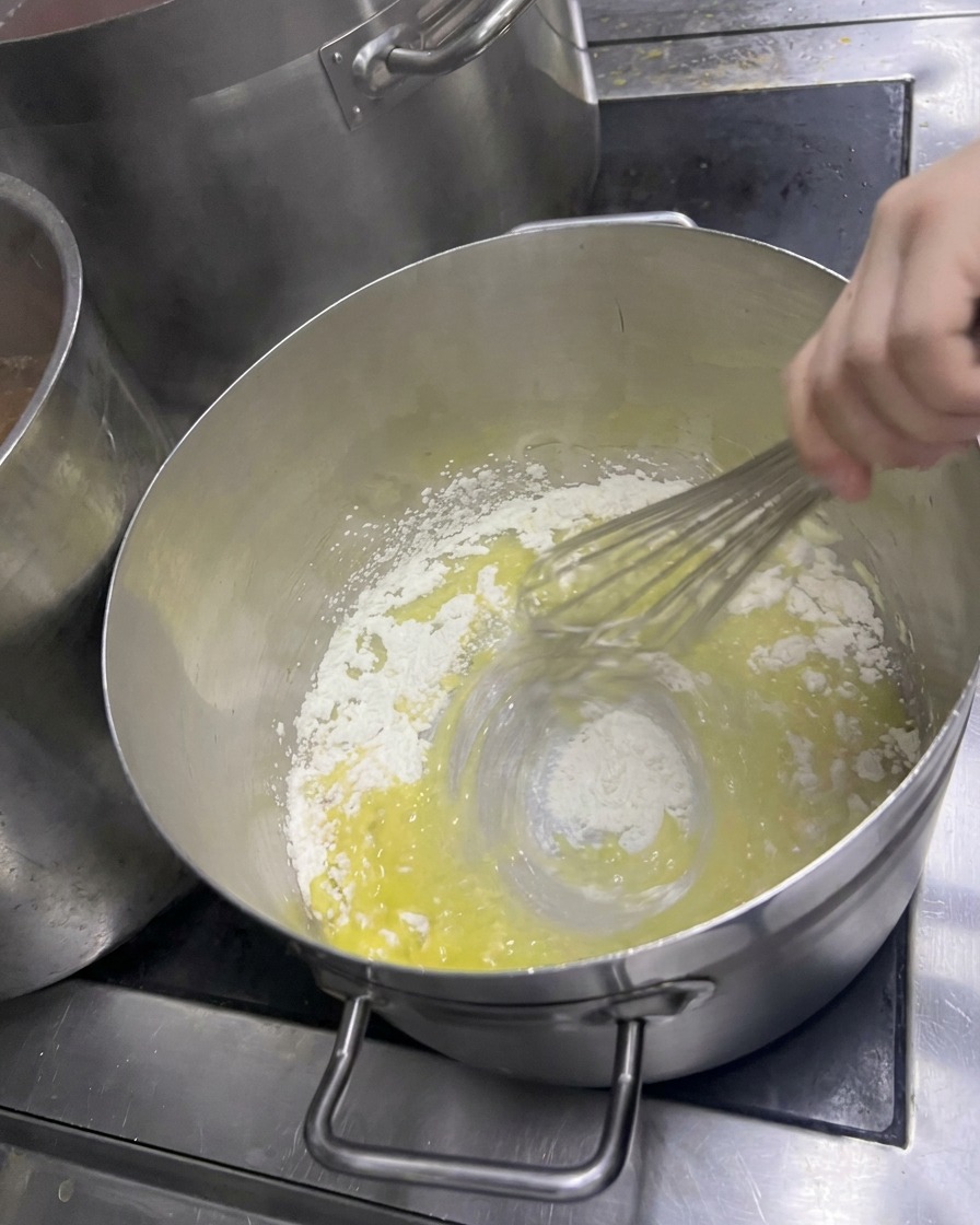 Una mano che mescola energicamente farina e burro fuso in una pentola per creare un roux giallo chiaro.