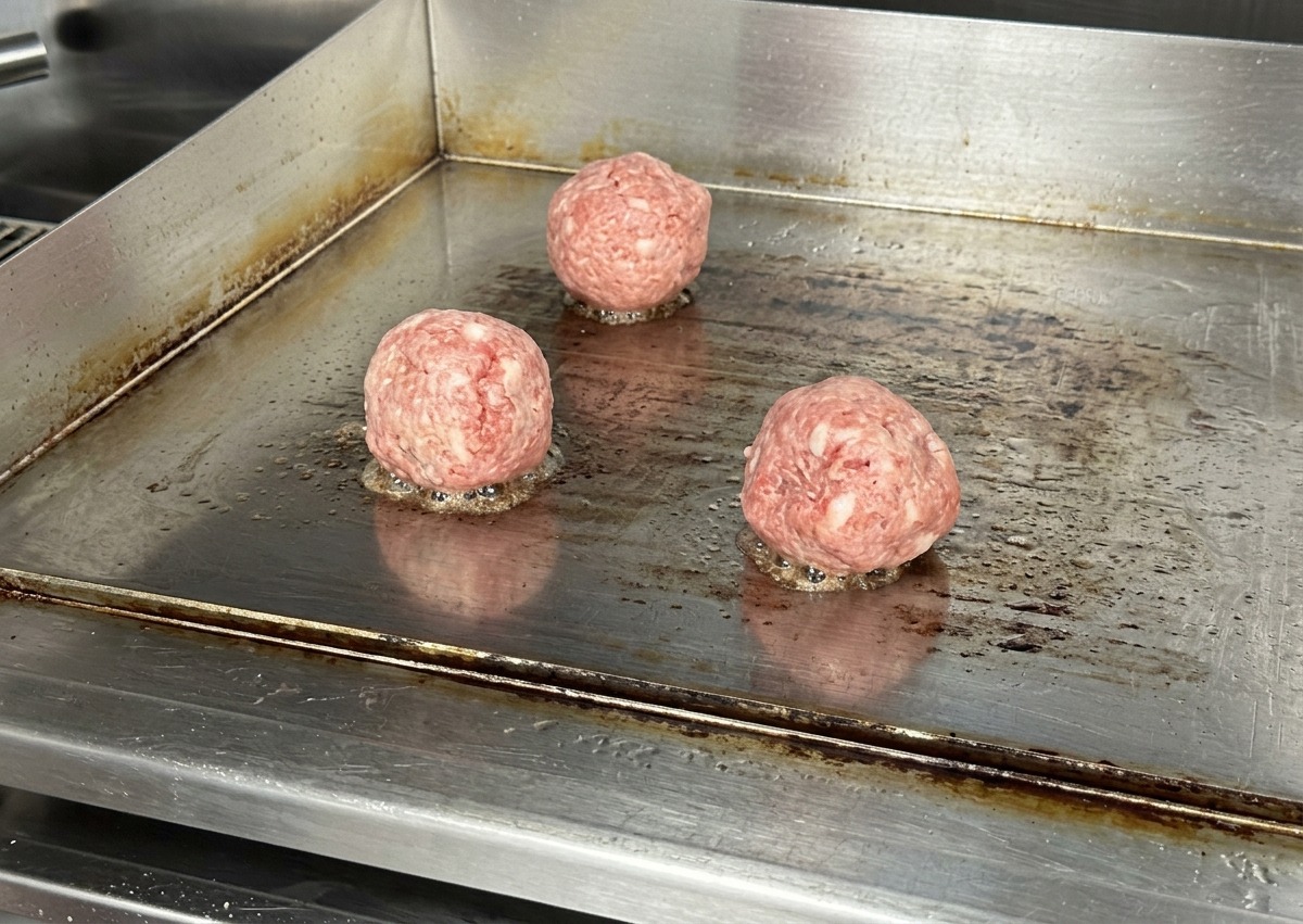 Tre palline tonde di carne di manzo cruda su una piastra piatta in acciaio inossidabile rovente.