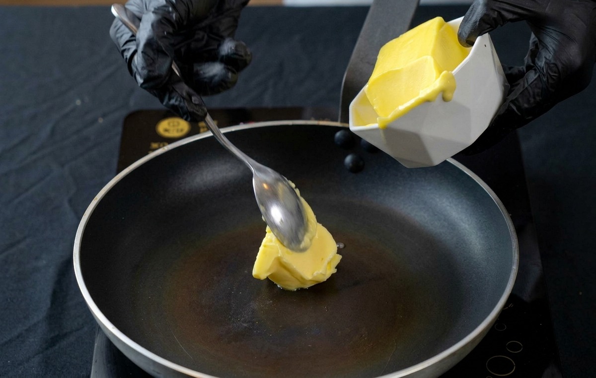 Una mano che aggiunge un pezzetto di burro giallo in una padella antiaderente calda.