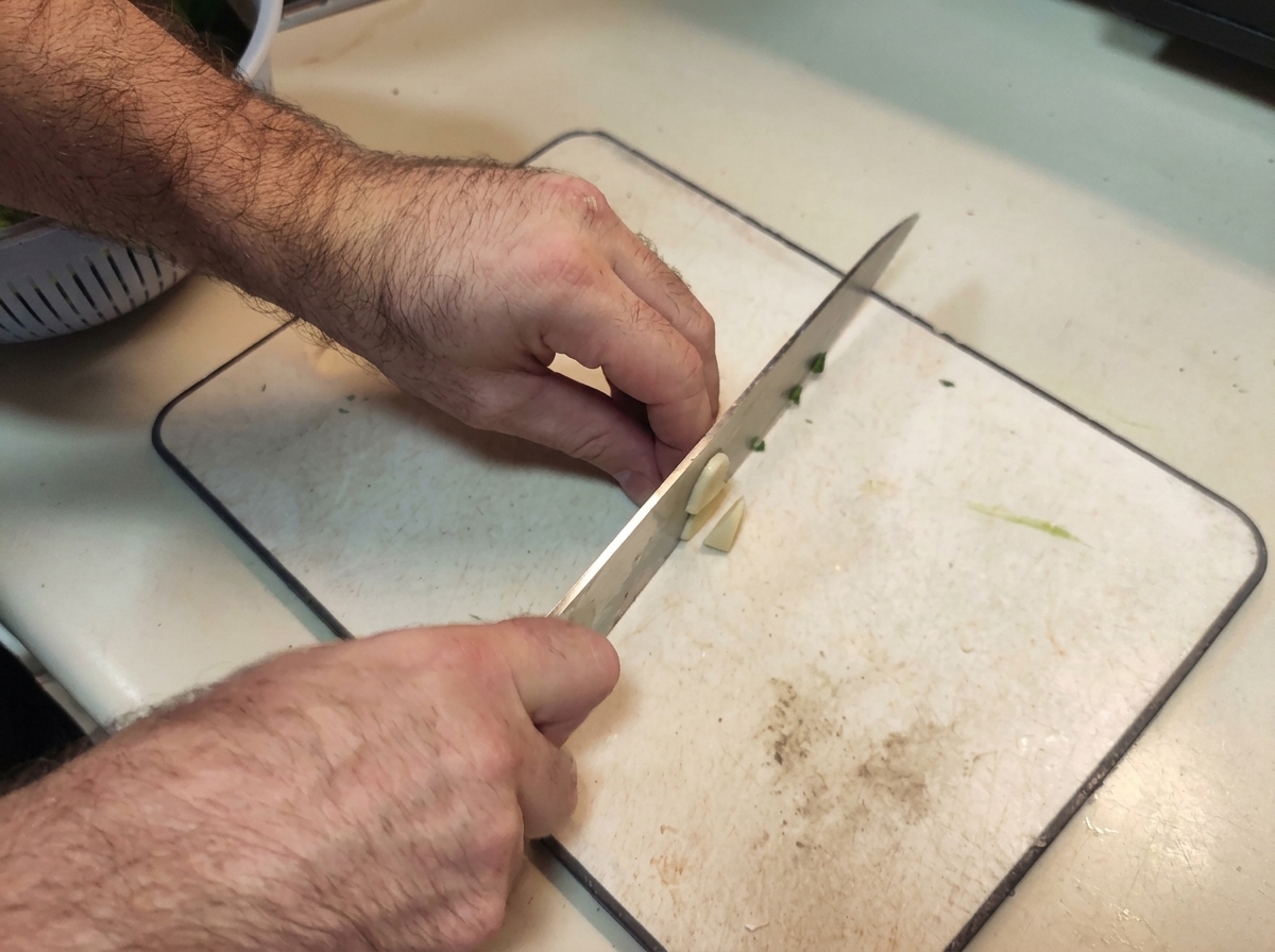 Mani che utilizzano un coltello da chef per tritare finemente spicchi daglio su un tagliere bianco.