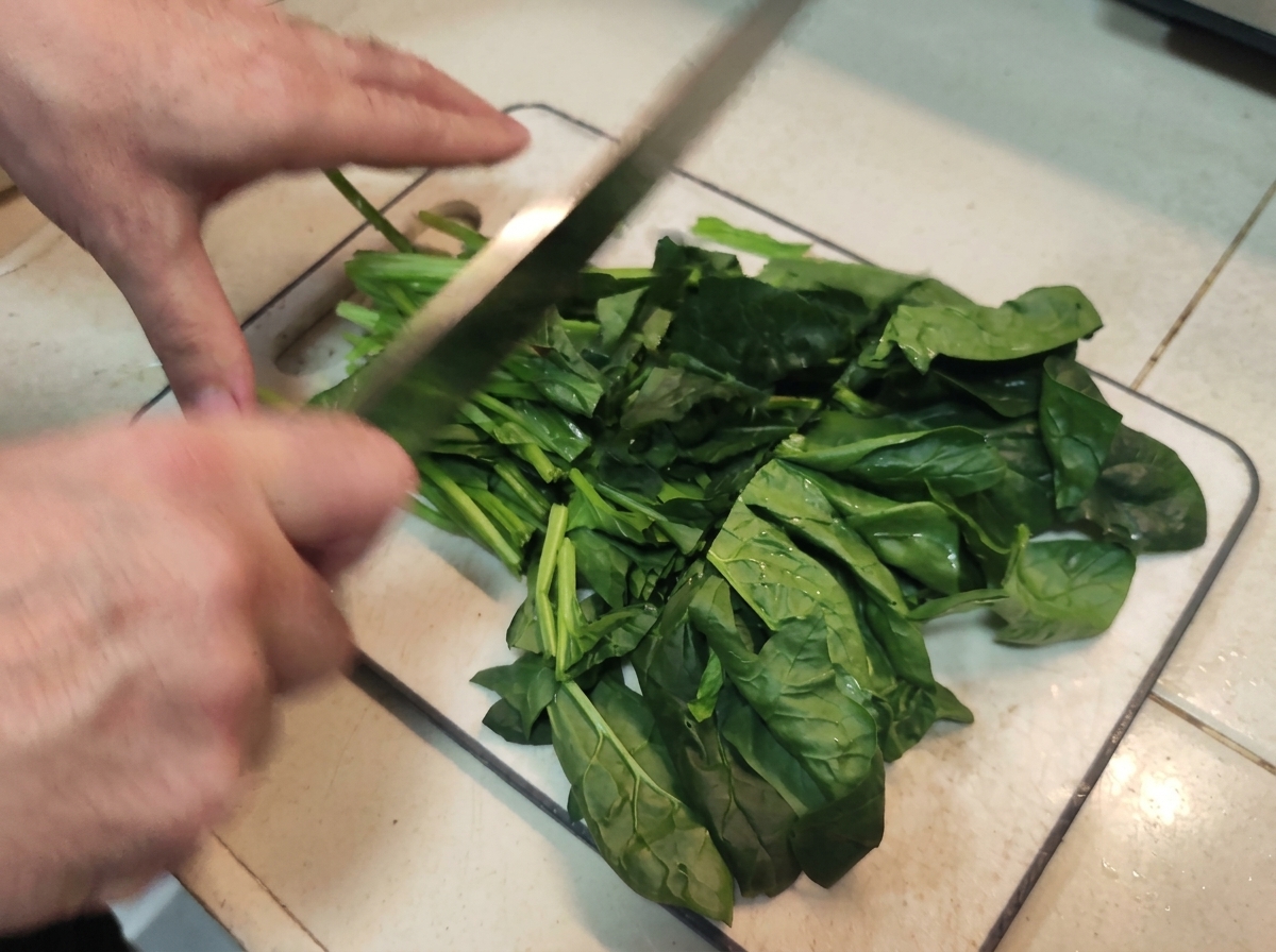 Mani che usano un grosso coltello per tagliare grossolanamente un mucchio di foglie di spinaci freschi.