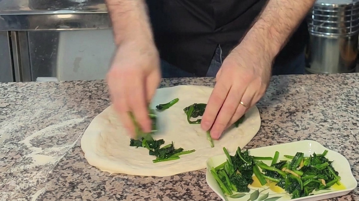 Mani che distribuiscono spinaci cotti su una base di pizza cruda stesa a mano su un piano infarinato.
