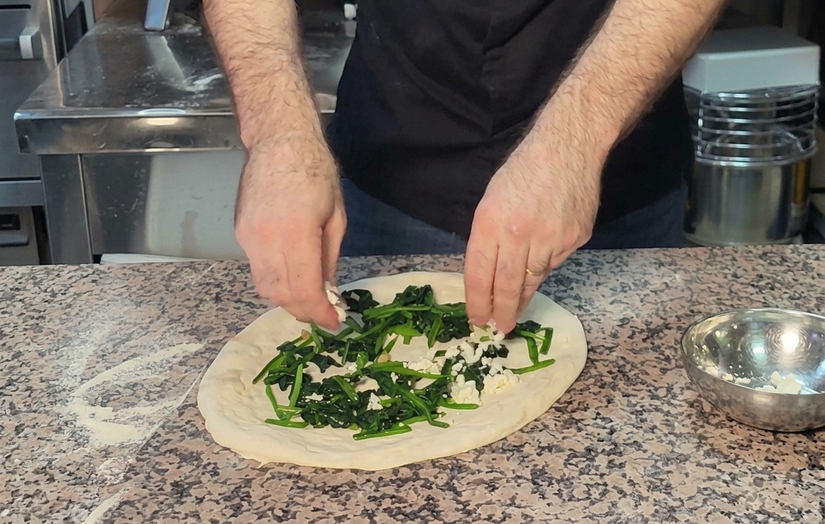 Mani che sbriciolano la feta sopra uno strato di spinaci verdi sullimpasto della pizza.