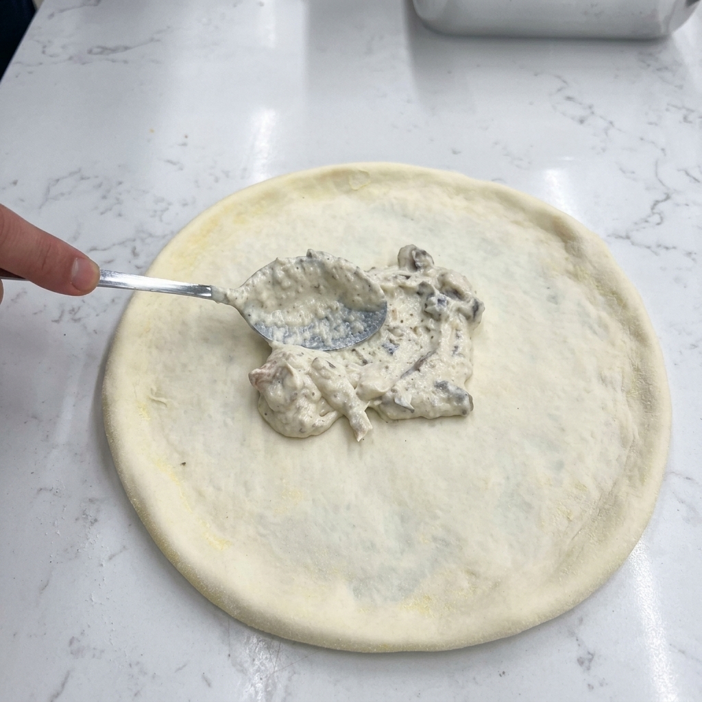 Una mano che usa un cucchiaio di metallo per spalmare la crema di funghi bianca sulla base della pizza.