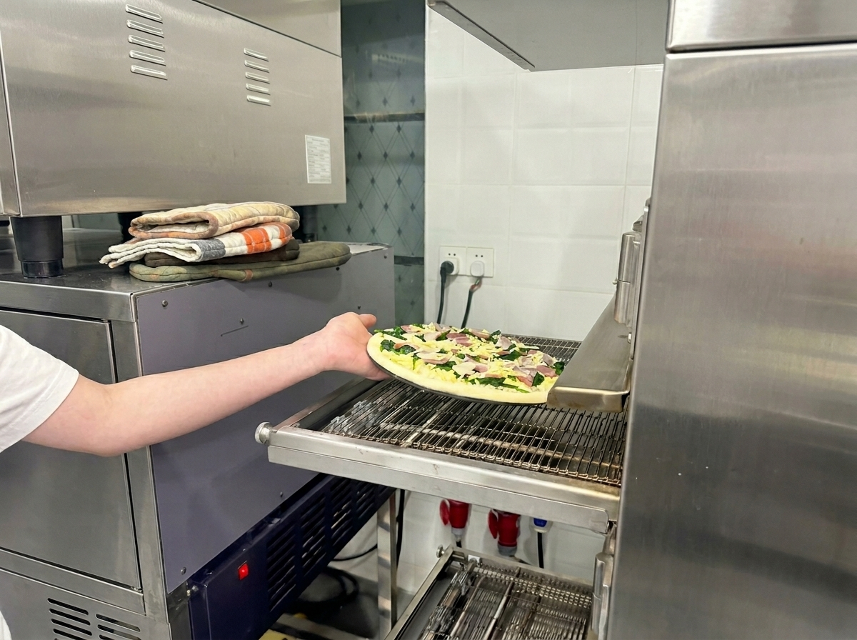Un braccio che posiziona la pizza cruda sul nastro trasportatore di un forno per pizza professionale.