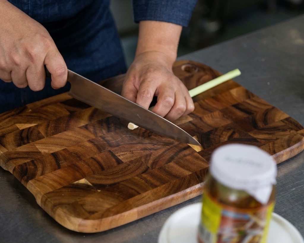 Mani che usano un coltello affilato per tritare finemente i gambi di citronella fresca su un tagliere di legno.