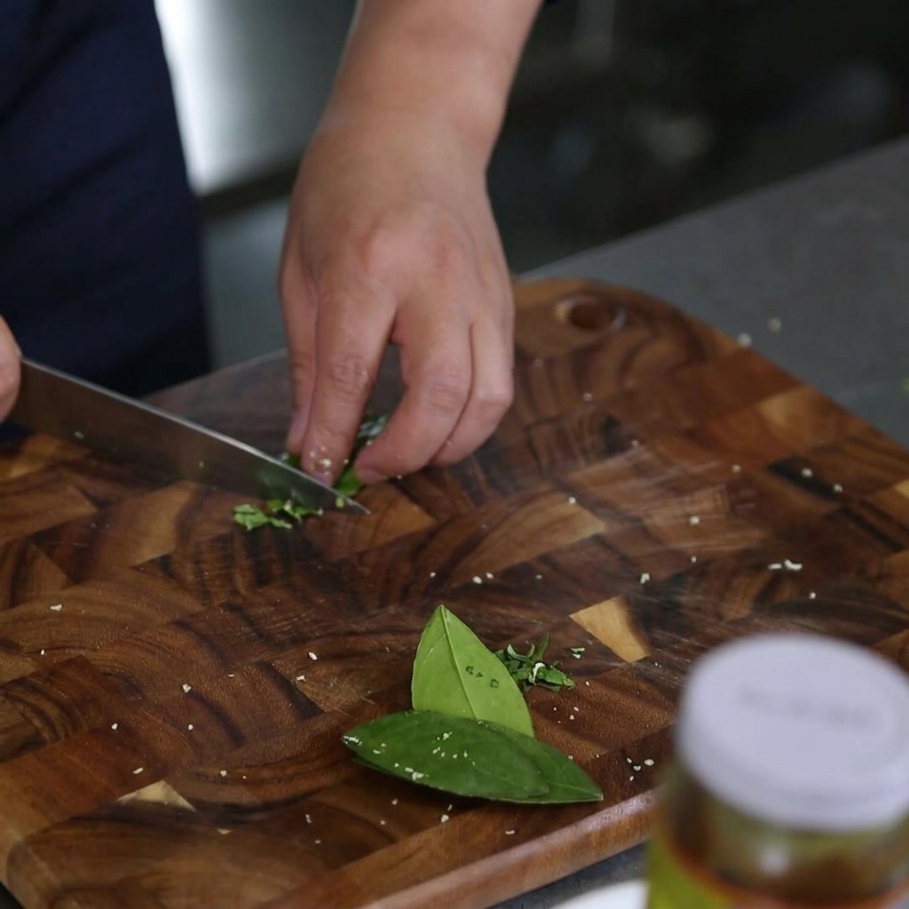 Primo piano di mani che usano un coltello da chef per rimuovere la venatura centrale dalle foglie di kaffir lime su un tagliere.