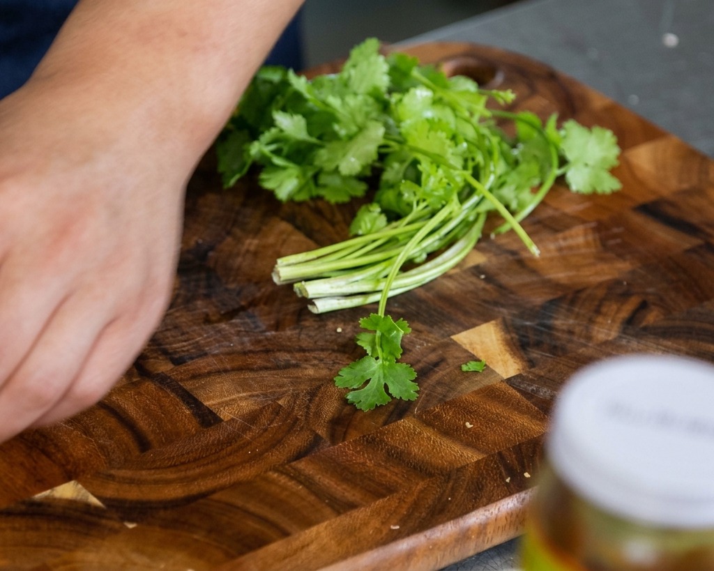 Foglie e gambi di coriandolo fresco su un tagliere di legno, pronti per essere sminuzzati a mano.