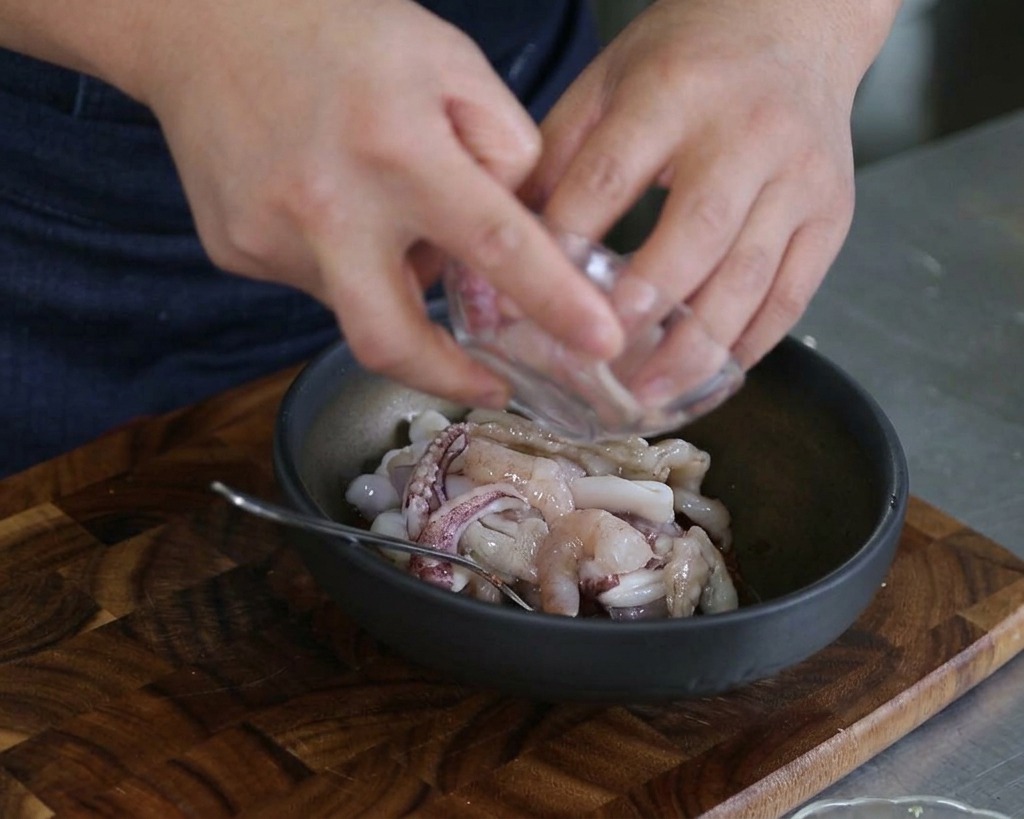 Mani che versano calamari a fette e gamberi crudi in una ciotola scura contenente salsa e altri frutti di mare.
