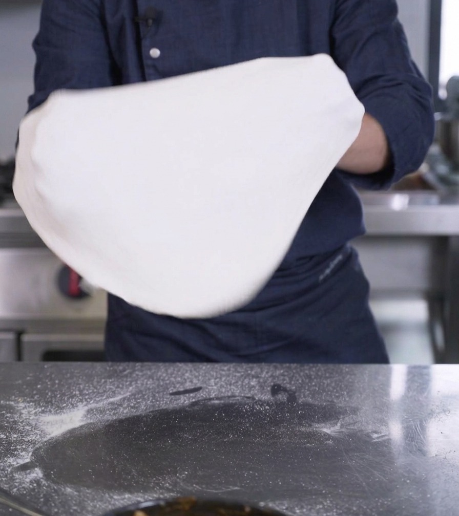 Mani di una persona che sollevano e stendono un grande disco di impasto per pizza bianco.