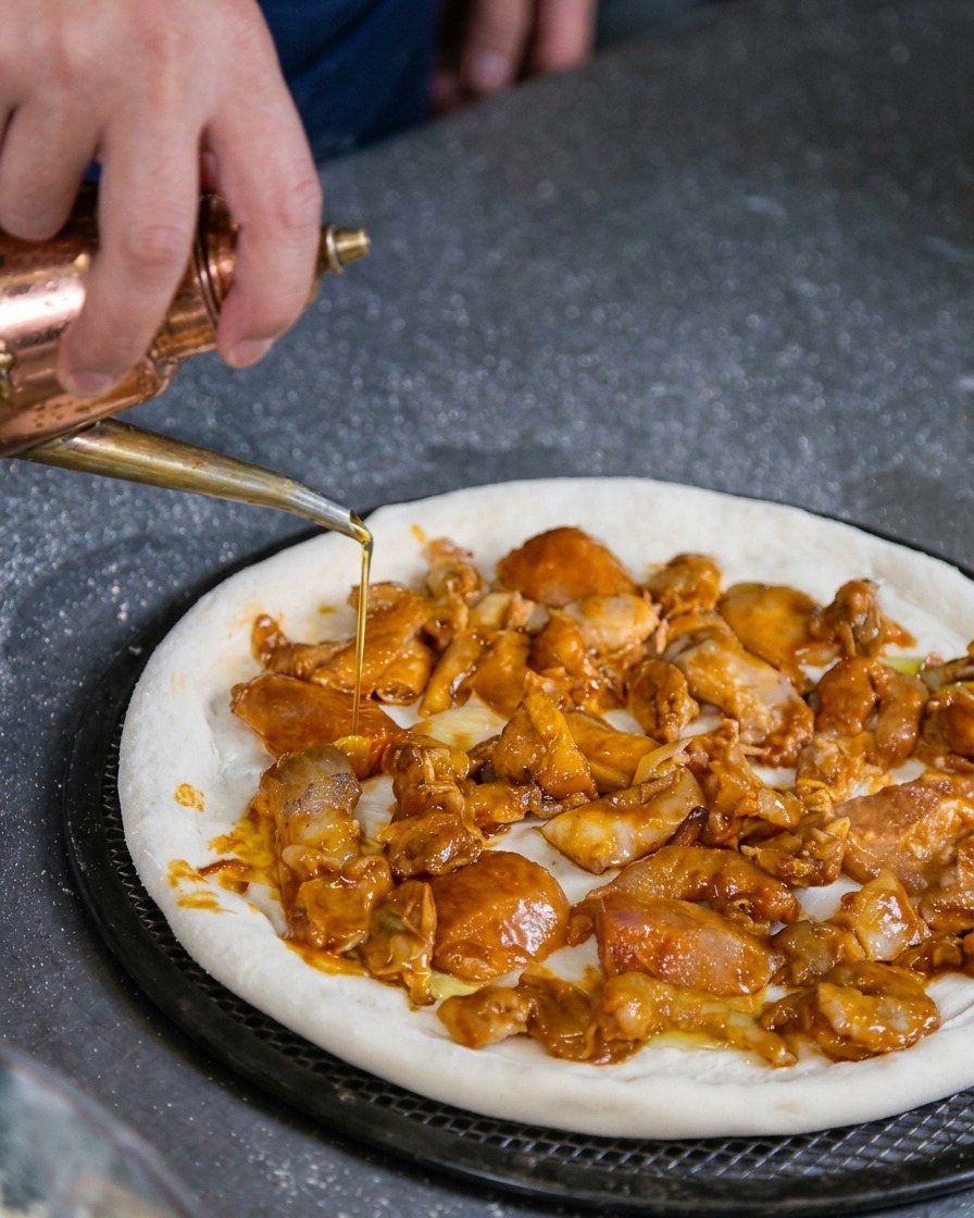 Una mano che usa un oliatore in rame per versare delicatamente olio doliva su una base pizza cruda coperta di frutti di mare.