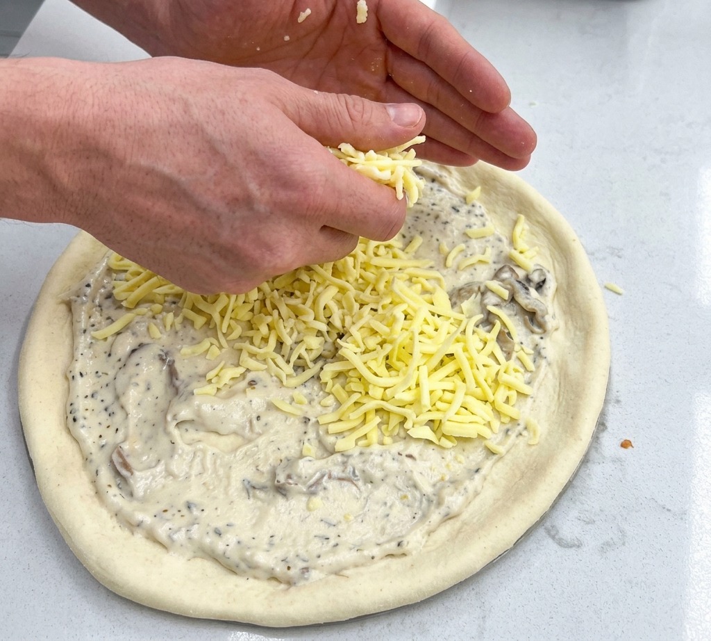 Mani che cospargono mozzarella grattugiata su una base pizza già ricoperta di crema bianca.