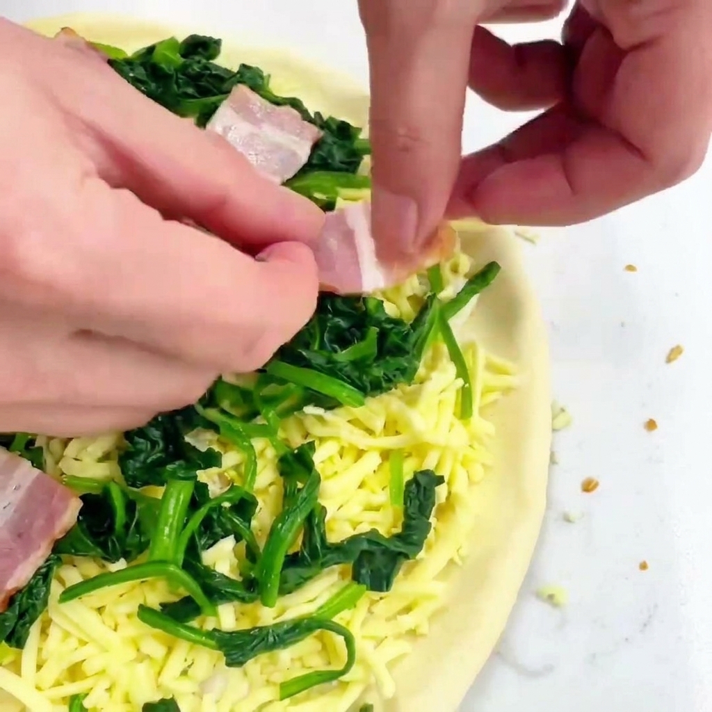 Mani che posizionano quadrati di pancetta cruda su una pizza con formaggio e spinaci.