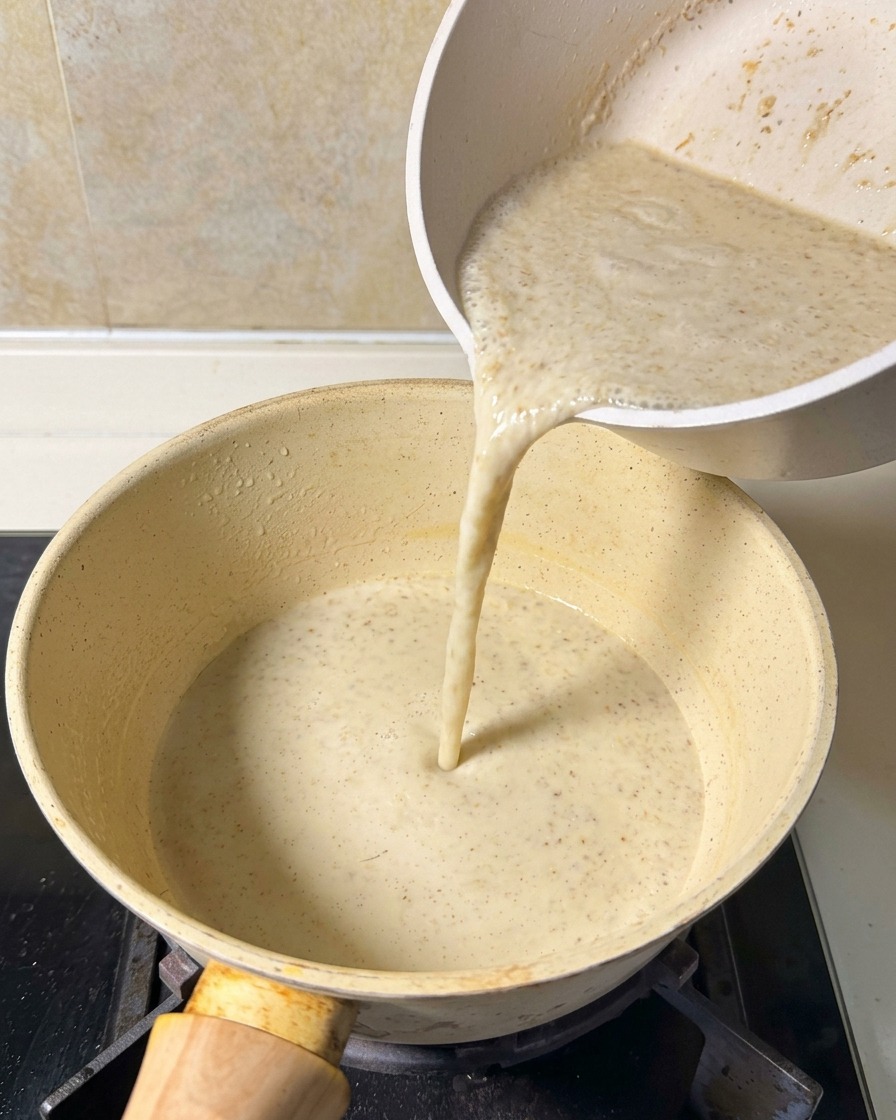 Un composto liscio e beige per la zuppa versato da una caraffa bianca in una pentola gialla sul fornello.