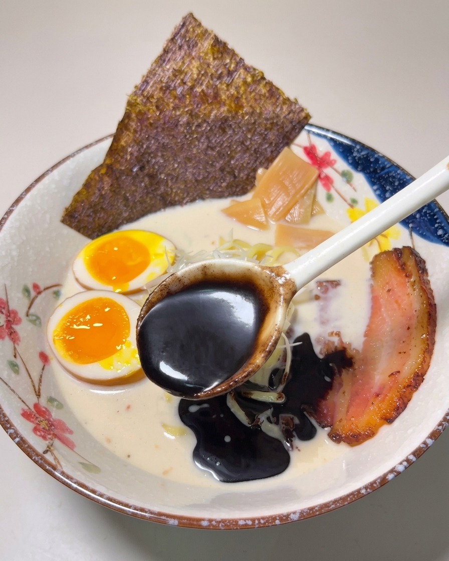 Un cucchiaio di olio allaglio nero, denso e scuro, versato sopra una ciotola di ramen finito.