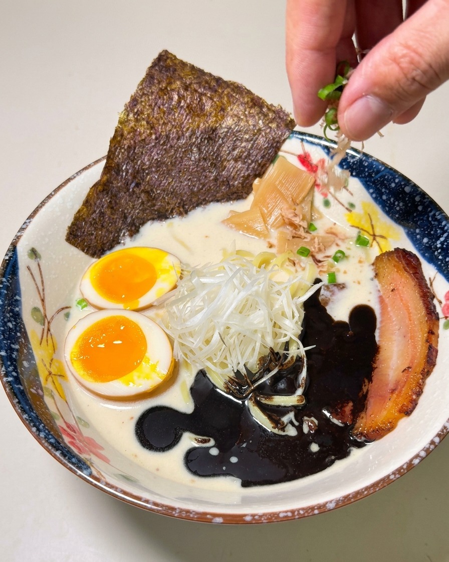 Una mano spolvera cipollotti tritati su una ciotola di ramen con olio allaglio nero, chashu e uovo.