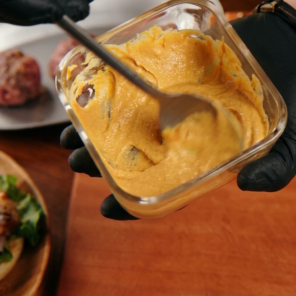 Una mano che mescola una salsa burger arancione e cremosa con un cucchiaio in un contenitore di vetro.
