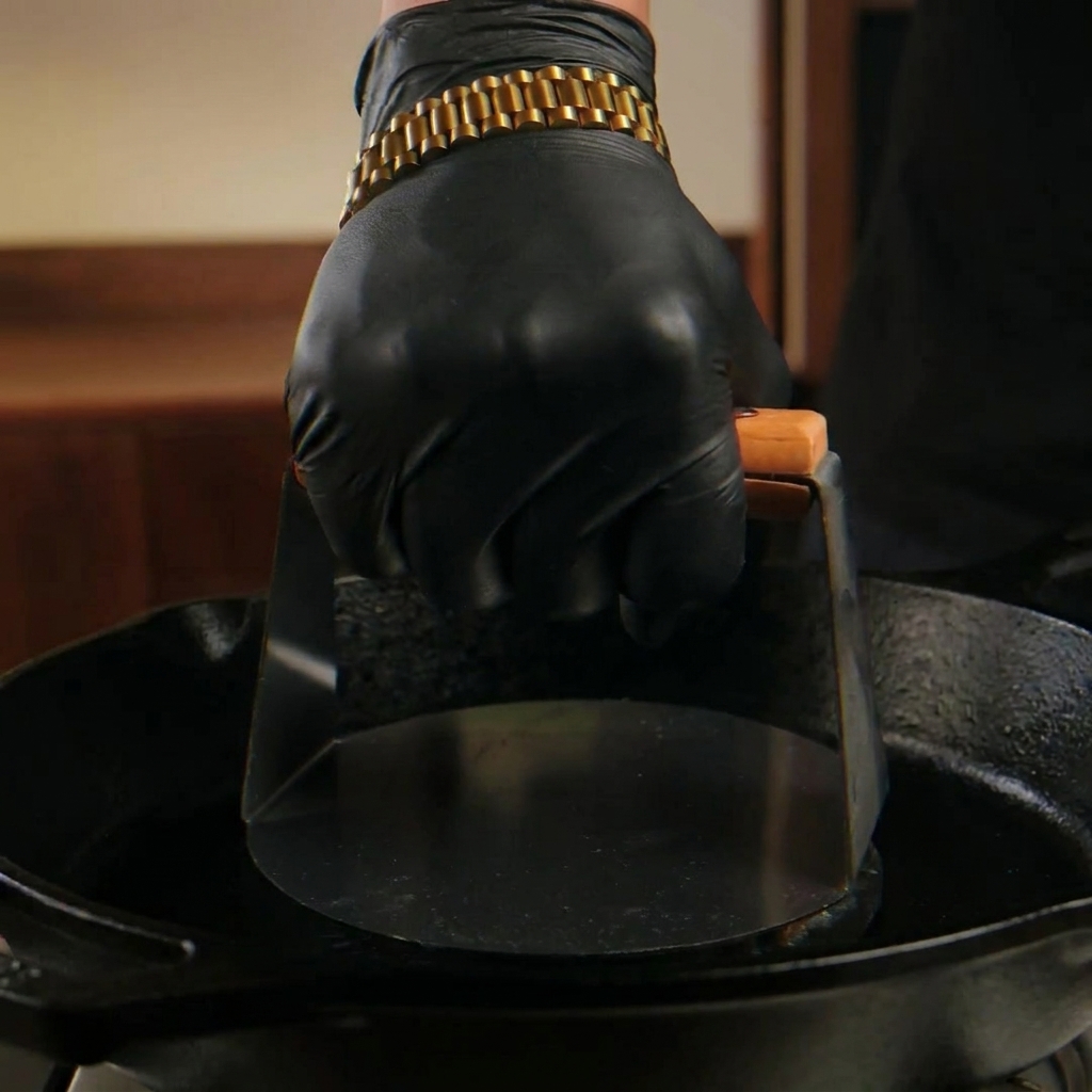 Una mano con guanto nero usa una pressa metallica pesante per schiacciare una pallina di manzo cruda su una padella di ghisa rovente.