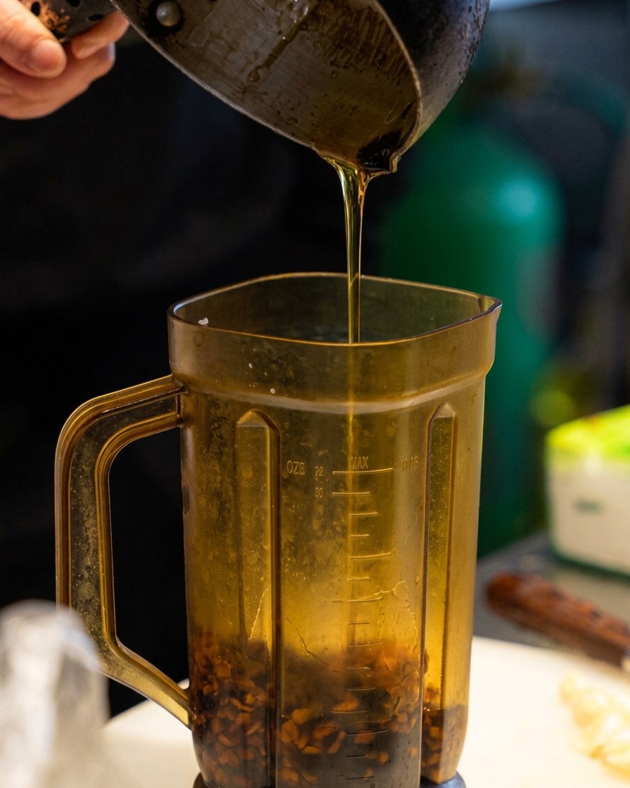 Olio di sesamo dorato versato da un pentolino in un frullatore contenente pezzetti di aglio scuro.