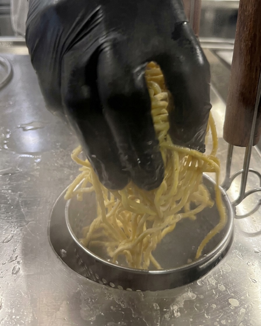 Una mano immerge una porzione di noodles gialli in una pentola di acqua bollente.