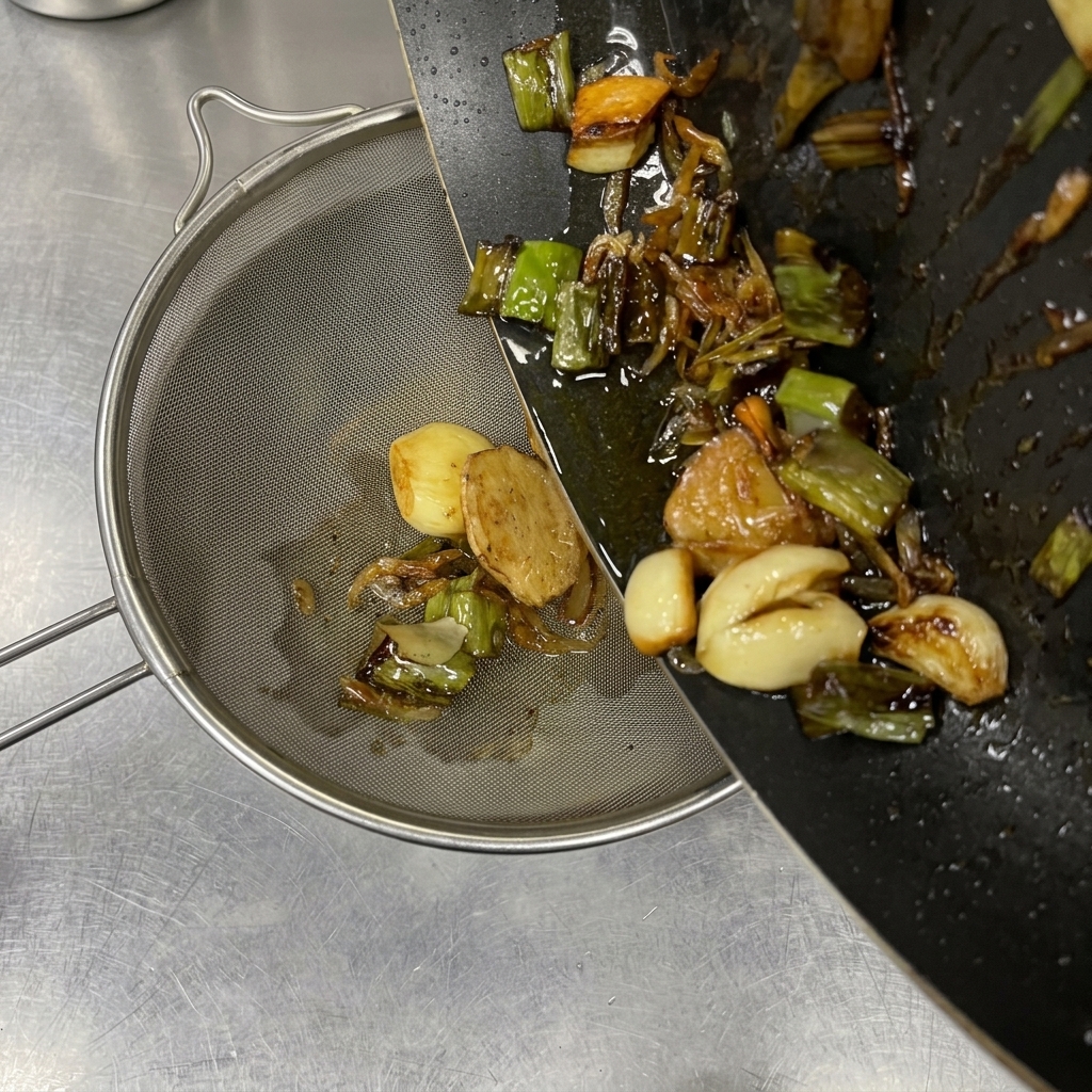 Olio caldo aromatizzato con aglio e cipollotti dorati, versato da una padella scura in un colino a maglie fini.
