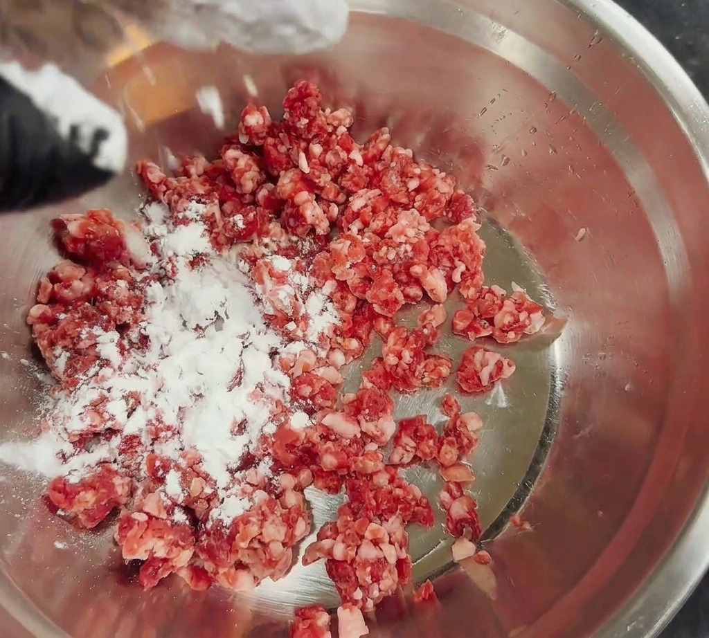 Amido di mais bianco spolverato sopra la carne macinata fresca in una ciotola di acciaio inox.