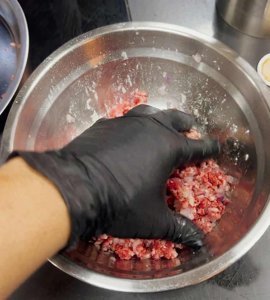 Una mano con guanto nero che mescola leggermente carne macinata e cipolle tritate in una ciotola dargento.