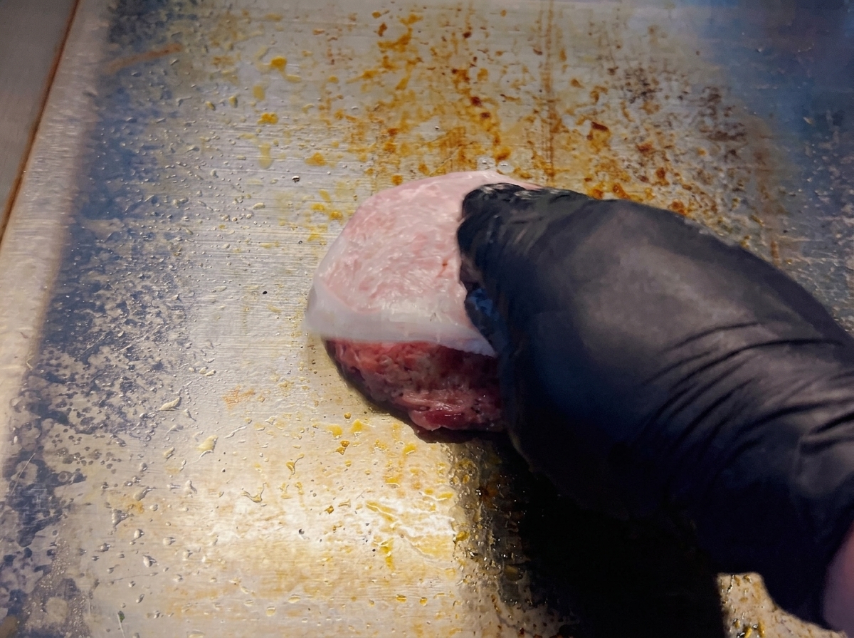 Una mano che rimuove la carta forno da un hamburger tondo posizionato su una piastra rovente.
