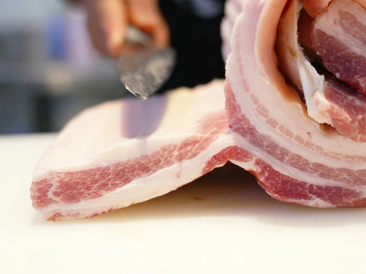 Un coltello affilato che incide un pezzo di pancetta di maiale cruda su un tagliere bianco.