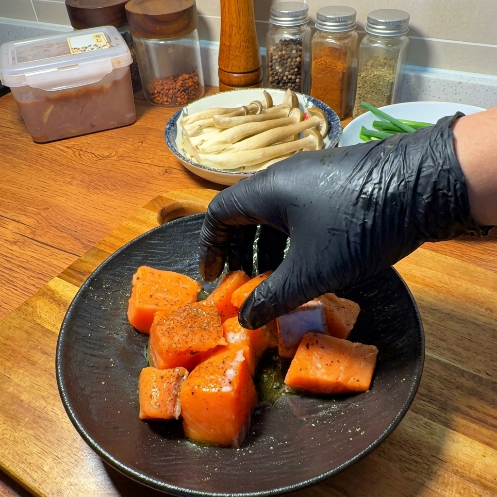 Una mano con guanto che mescola i cubetti di salmone con il condimento e lolio doliva su un piatto scuro.