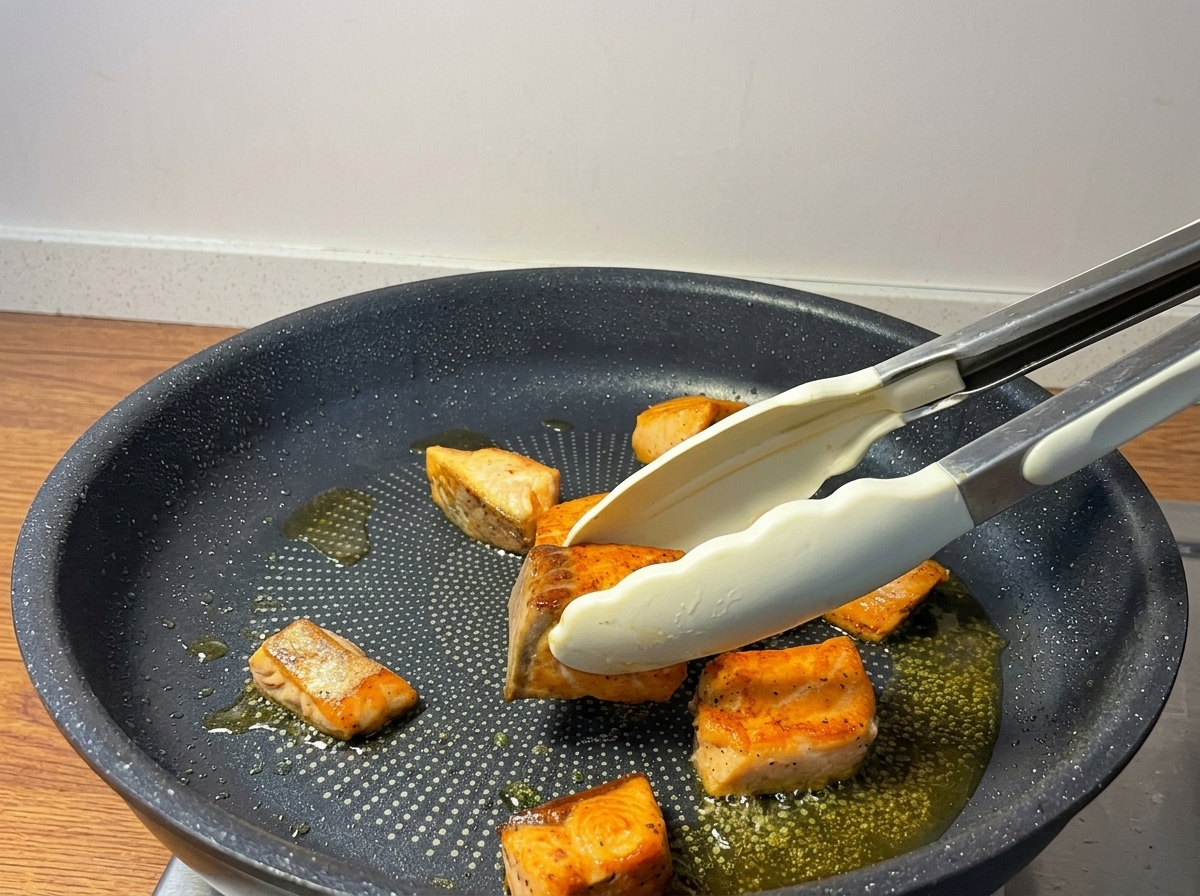Pinze da cucina che girano cubetti di salmone dorati in una padella antiaderente.