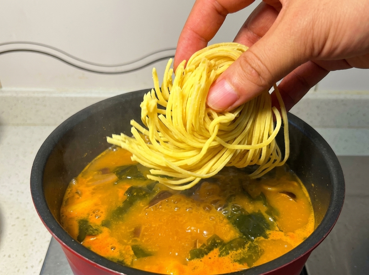 Una mano che immerge un nido di noodle gialli per ramen in una pentola di brodo arancione.