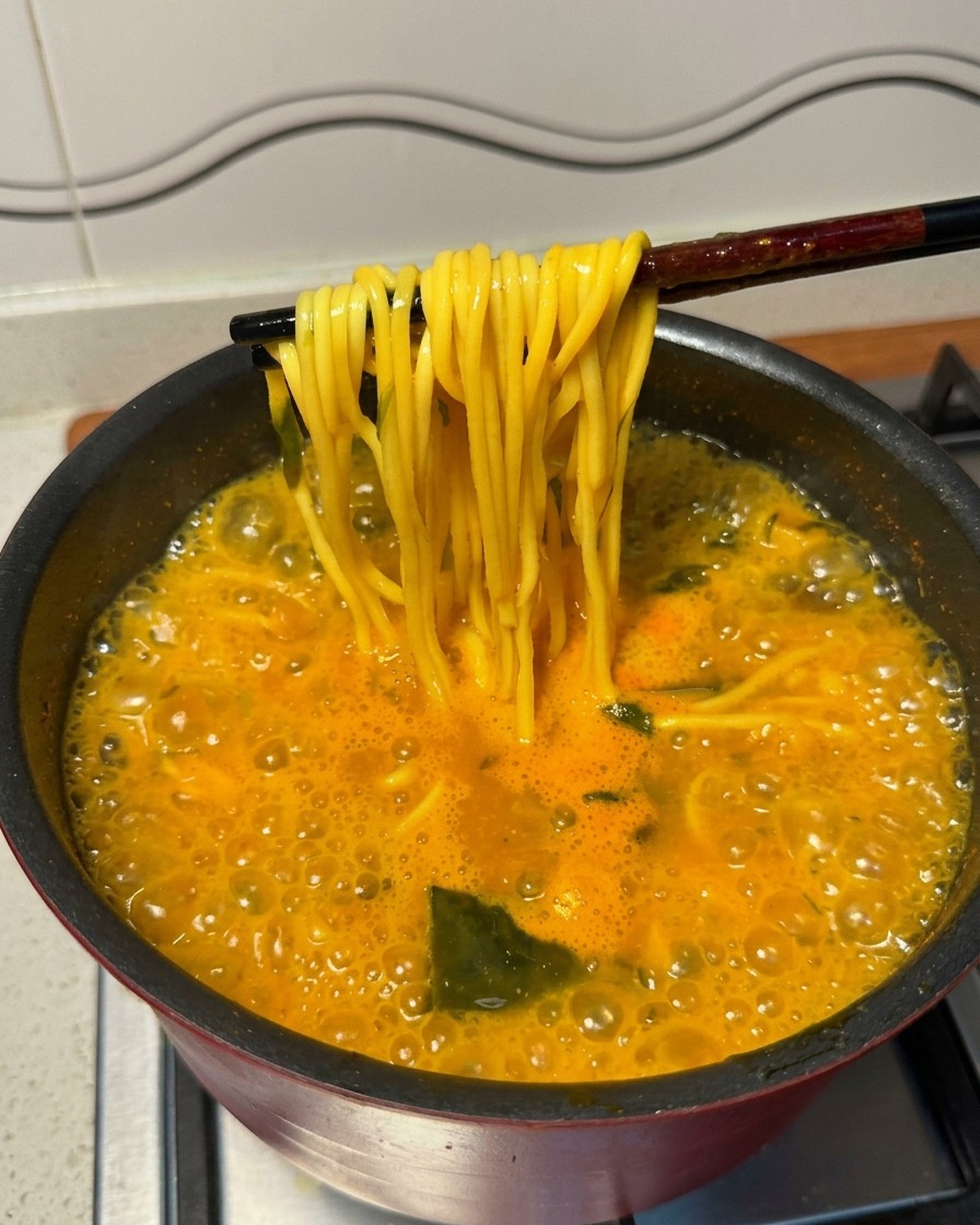 Bacchette nere che sollevano i noodle gialli cotti da una pentola fumante di zuppa piccante.