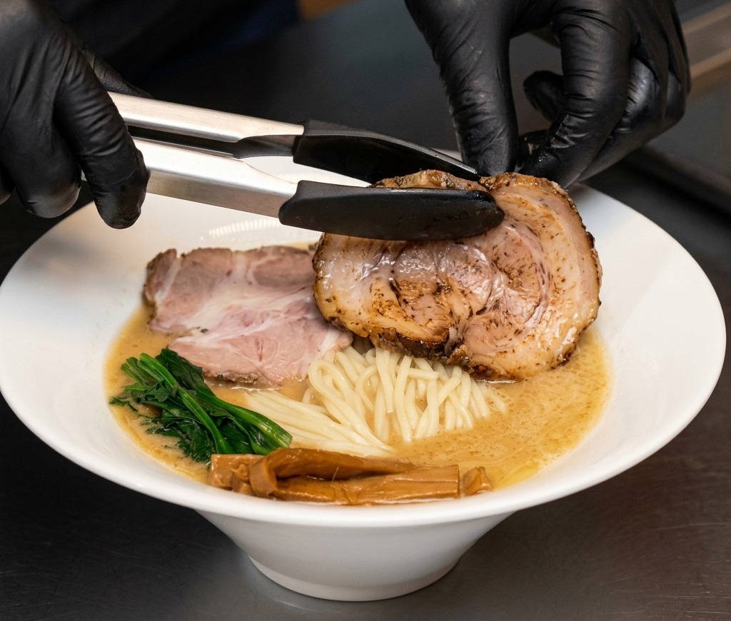 Pinze da cucina che adagiano una grande fetta di pancetta chashu in una ciotola di ramen.
