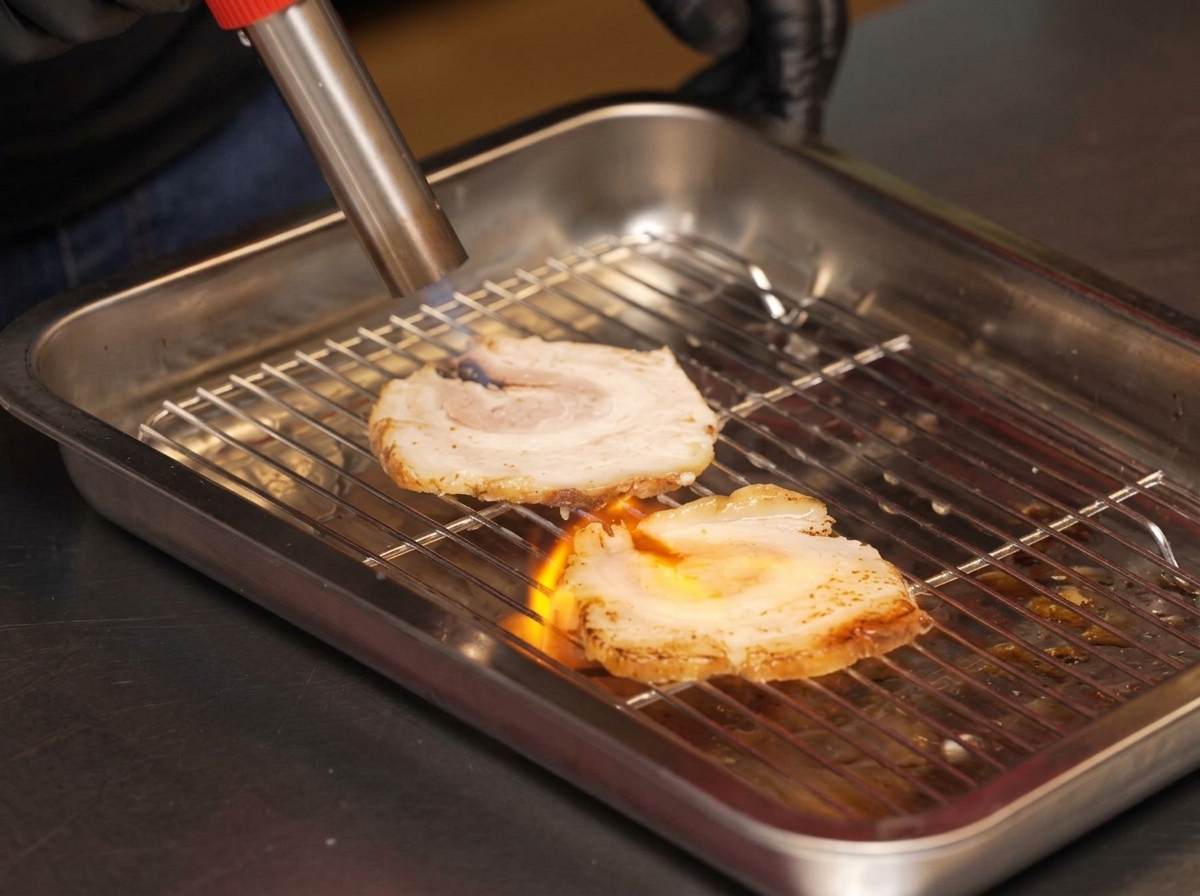 Uno chef usa un cannello da cucina per scottare le fette di maiale chashu su una griglia.