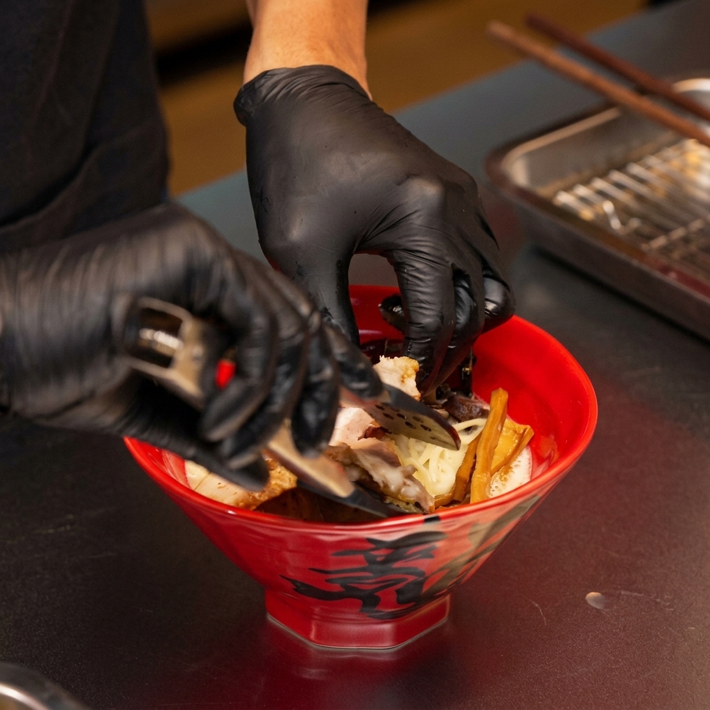 Mani che usano pinze per adagiare le fette di maiale chashu scottate in una ciotola di miso ramen.
