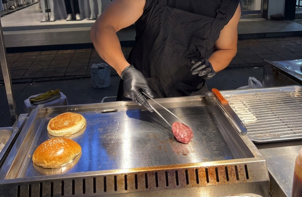 Una persona che posiziona una pallina di carne macinata cruda su una piastra di metallo rovente e fumante.