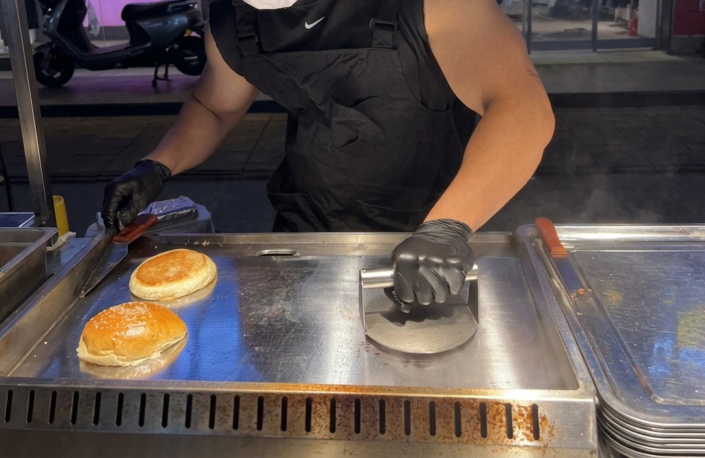 Una persona che usa una pressa pesante in metallo per appiattire con forza una pallina di carne sulla piastra.