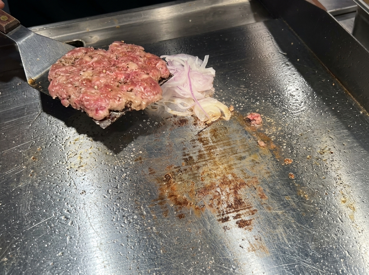 Una spatola di metallo che gira un burger ben rosolato sopra il letto di cipolle sulla piastra.