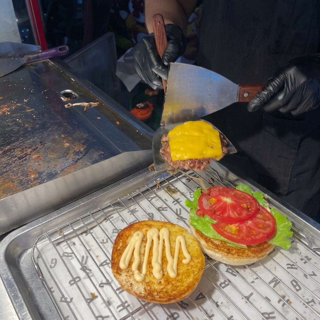 Una persona che usa una spatola per mettere il patty cotto con formaggio fuso sopra la base del burger con lattuga e pomodoro.
