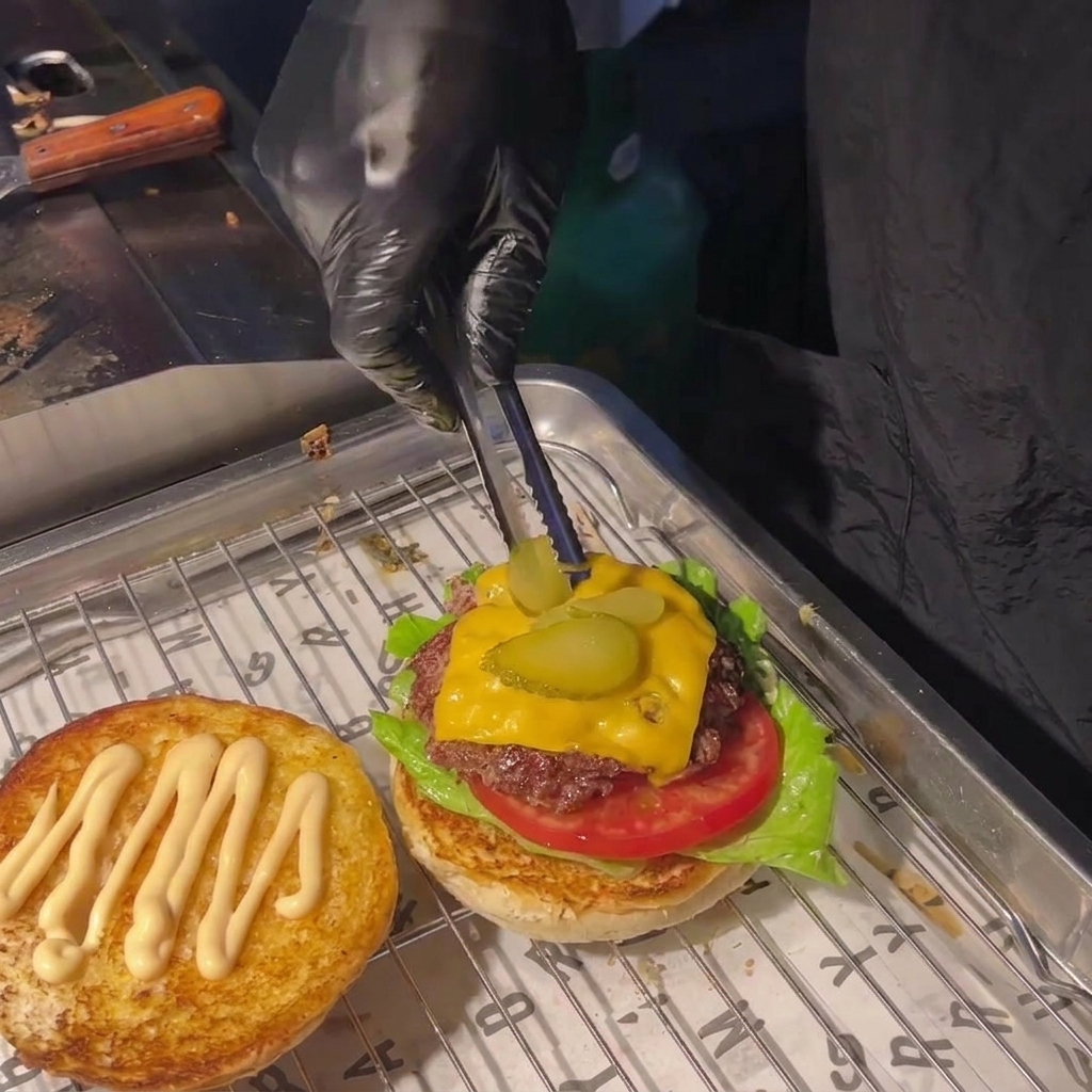 Una persona che usa delle pinze per mettere due fette di cetriolino sopra il formaggio fuso del burger.