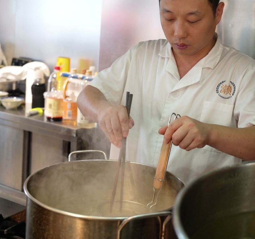 Uno chef usa bacchette lunghe di legno per girare i noodles freschi in un colino a rete metallica immerso in una pentola di acqua bollente.