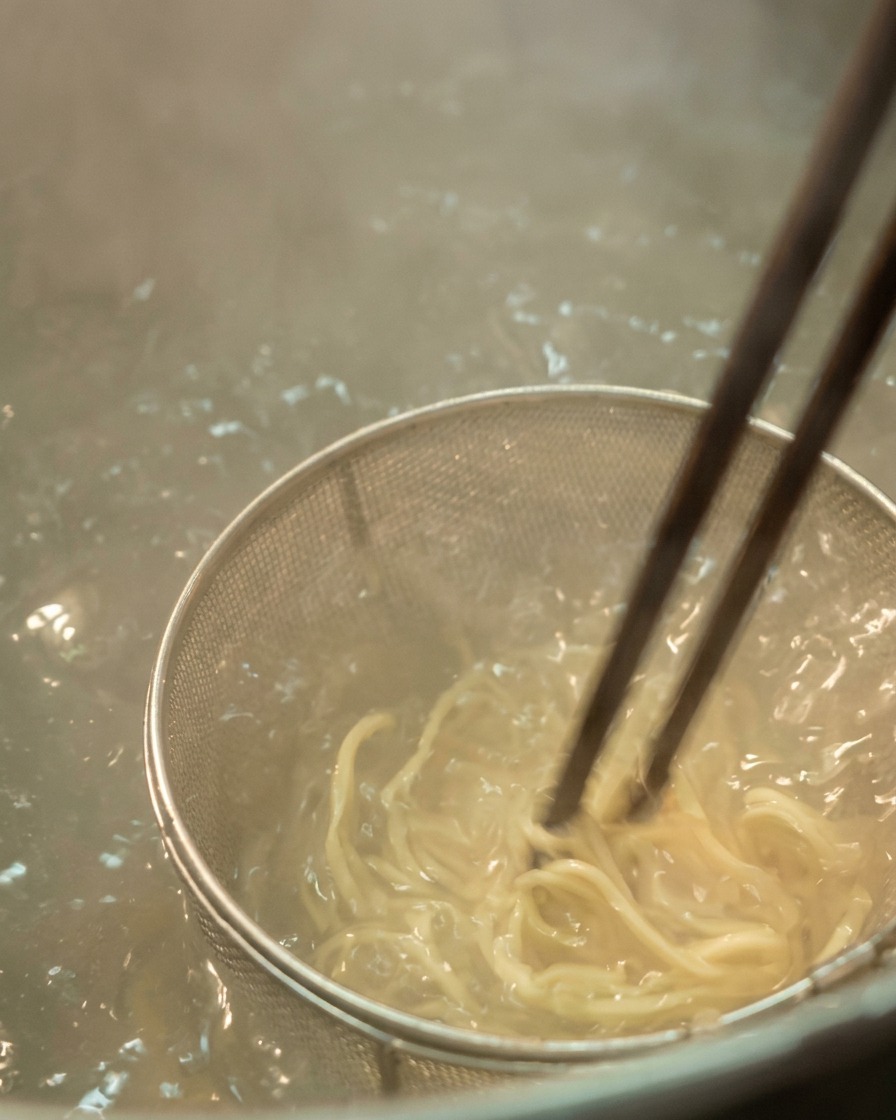 Dettaglio di noodles chiari mescolati con bacchette scure in un colino a rete metallica immerso in acqua bollente gorgogliante.