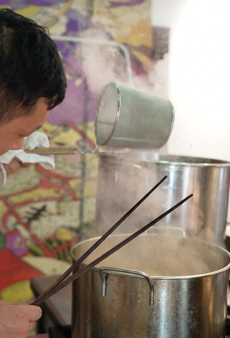 Uno chef scuote un colino cilindrico per noodles sopra una pentola fumante per eliminare lacqua in eccesso.